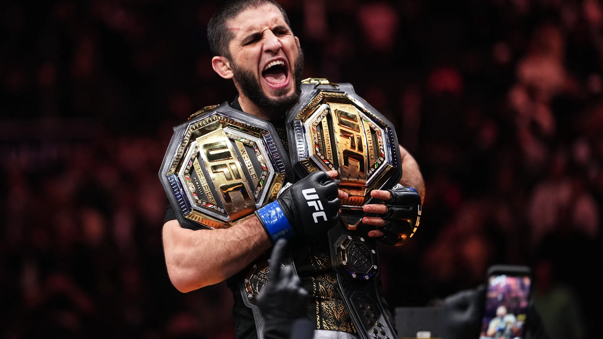 Islam Makhachev roaring while holding two UFC championship belts