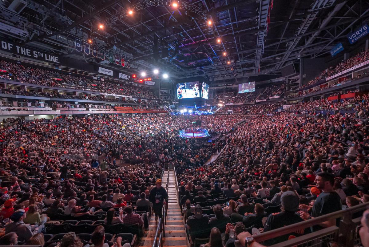A packed UFC arena with the octagon centered under bright lights and a massive overhead screen
